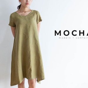 May include: A loose-fitting, olive green linen dress with a v-neck and short sleeves. The dress has a slightly asymmetrical hemline. The dress is made by the brand "MOCHA modern + comfort".