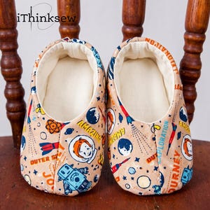 A pair of handmade soft-sole baby shoes featuring a retro outer space and astronaut fabric print, sitting on a wooden chair. Represents the finished Abella Baby Shoes pattern.