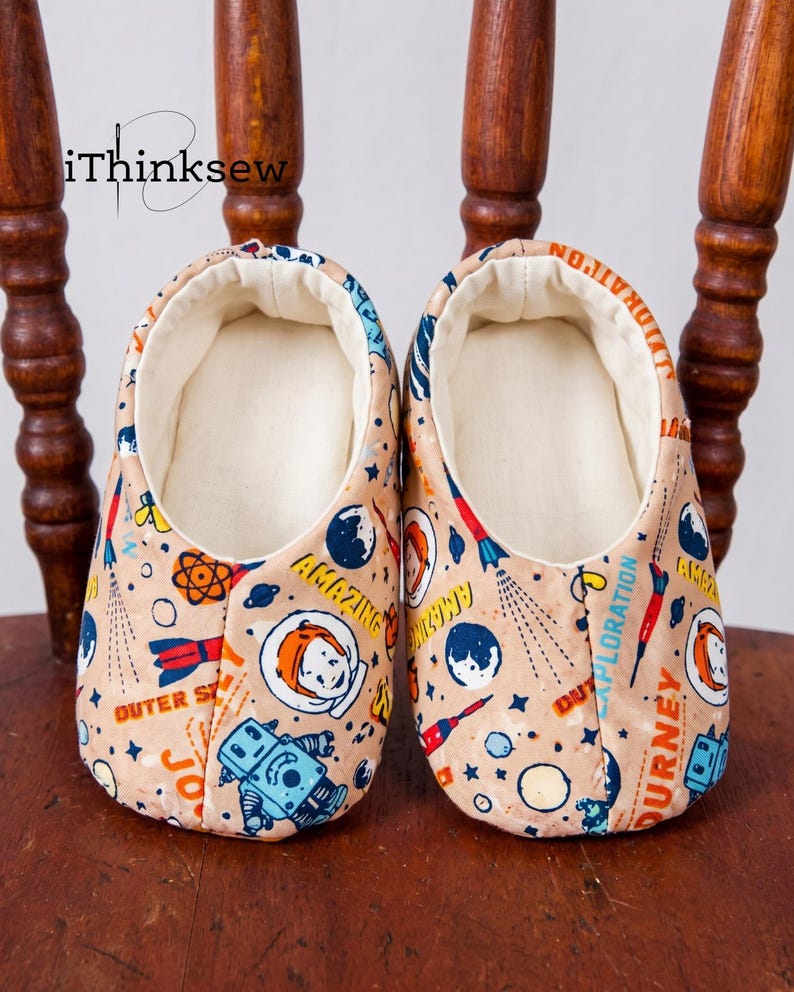 A pair of handmade soft-sole baby shoes featuring a retro outer space and astronaut fabric print, sitting on a wooden chair. Represents the finished Abella Baby Shoes pattern.