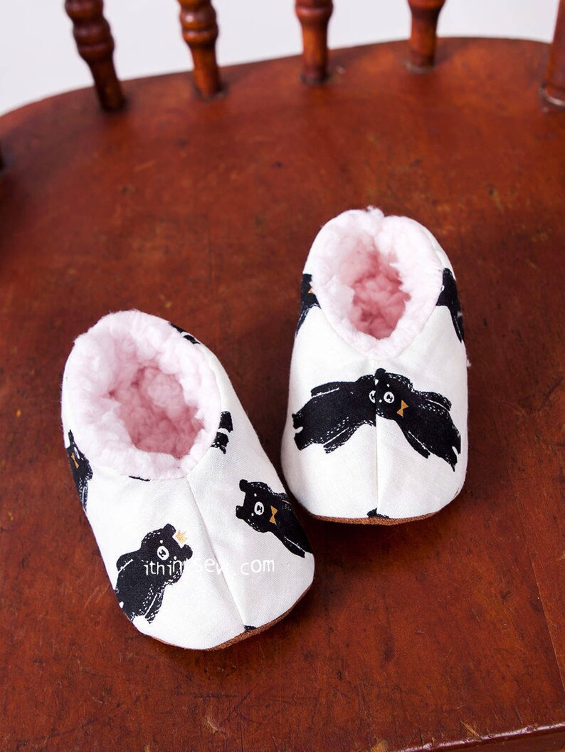 May include: A pair of white baby booties with a black bear print and pink fleece lining. The booties are sitting on a wooden chair.