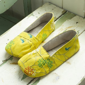 Op de afbeelding: Een paar gele stoffen slippers met een wit en groen bloemenpatroon. De slippers hebben een simpel ontwerp en zien er comfortabel uit.