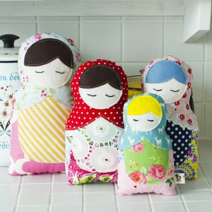May include: Five colorful fabric nesting dolls with embroidered faces and various patterns. The dolls are arranged in a row from largest to smallest. The largest doll is pink and yellow with a floral pattern. The second largest doll is red and white with polka dots. The third largest doll is blue and white with polka dots. The fourth largest doll is green and pink with floral patterns. The smallest doll is light blue with a deer pattern.