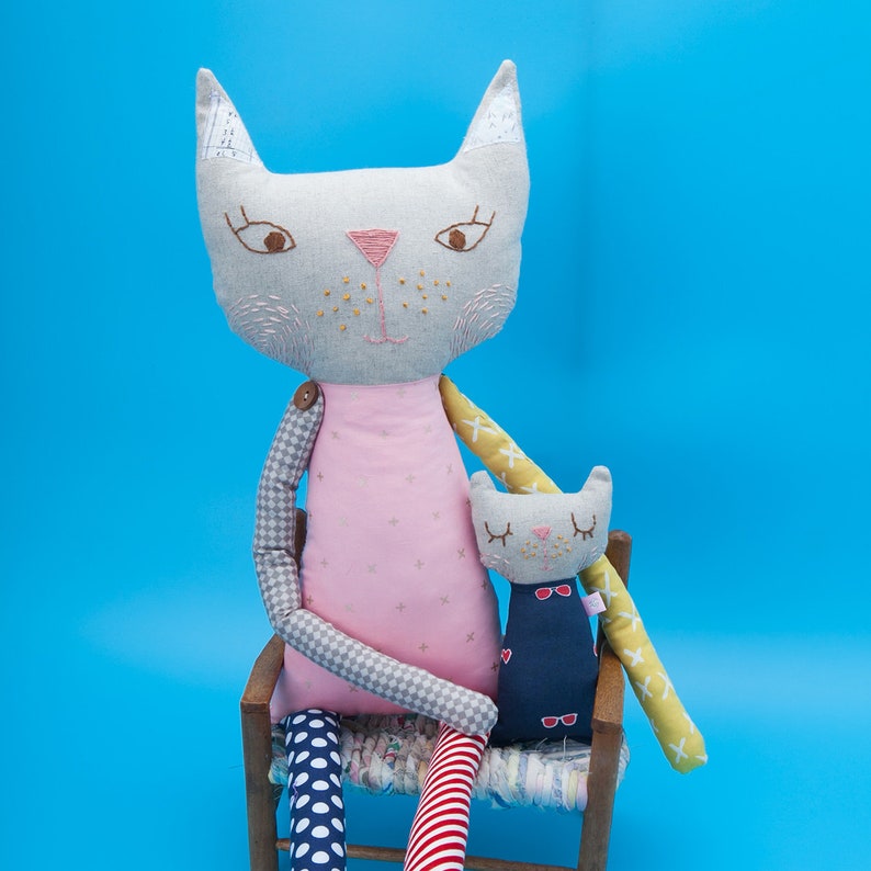 May include: Two gray fabric cat dolls sitting on a chair. The larger cat is wearing a pink dress and has a yellow and white striped arm. The smaller cat is wearing a blue and white striped dress and has a red and white striped arm.