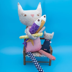 May include: A gray cat plush toy with embroidered details sits on a wooden chair holding two fish plush toys. The cat is wearing a pink dress with white polka dots and has a white and gray striped leg. The chair has a woven seat and a smaller gray cat plush toy with red and white striped legs is sitting on the chair.