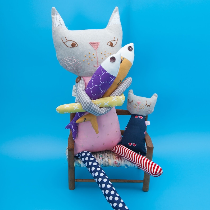 Whimsical handmade cat ragdoll with long patterned legs sitting on a wooden chair, holding two stuffed fish next to a smaller baby kitty. A colorful, DIY cloth doll sewing pattern on a bright blue background.