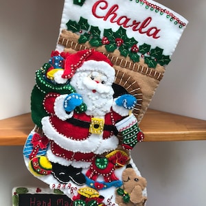 May include: A white felt Christmas stocking with a Santa Claus design. The stocking is decorated with beads, sequins, and embroidery. The stocking is personalized with the name "Charla".