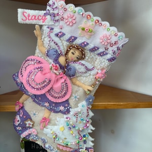 Könnte beinhalten: Ein weißer Weihnachtsstrumpf aus Filz mit dem in Pink gestickten Namen Stacy. Der Strumpf ist mit einer Fee in einem lila-rosa Kleid, Pailletten, Perlen, Stoffblumen und zwei Lebkuchenmännern am unteren Rand verziert.