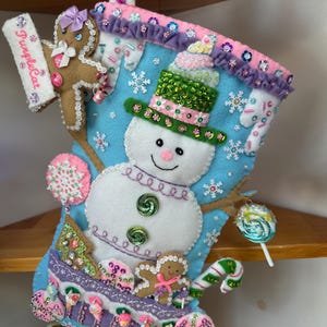 May include: A light blue felt Christmas stocking decorated with a central snowman wearing a green sequined hat, gingerbread figures, candy canes, and lollipops. The stocking features pink and purple trim, white snowflake accents, and sequins.