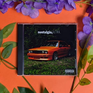 以下が含まれることがあります： アルバム「nostalgia, ULTRA.」のCDケース。カバーアートは、草むらに駐車されたオレンジ色の車を描いています。「nostalgia, ULTRA.」の文字が上部にあります。ケースは、オレンジ色の背景に紫色の花と緑の葉で囲まれています。