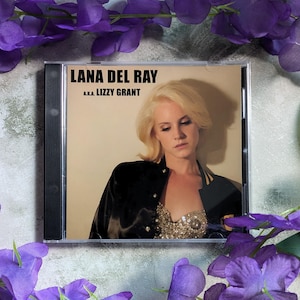 May include: A CD case featuring Lana Del Rey's album cover. The album title is displayed in bold black text, with "a.k.a. Lizzy Grant" below. The cover art shows a woman with blonde hair wearing a black jacket and a sequined top. Purple flowers surround the CD case.