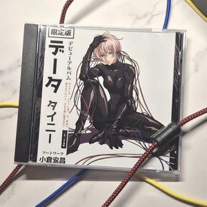 Può includere: Una custodia per CD con testo giapponese e un'illustrazione di una persona in tuta nera con capelli rosa, avvolta in fili. La custodia del CD è nera e trasparente. Cavi rossi, blu e gialli in primo piano.