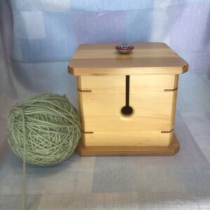 May include: A wooden yarn bowl with a single hole in the top and a small, colorful ceramic knob on the lid. A ball of green yarn sits next to the bowl.