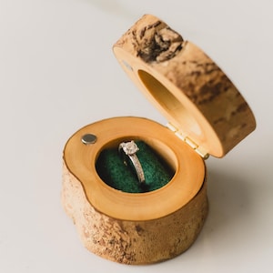 May include: A wooden ring box, made from a section of a tree branch, with a hinged lid. Inside, a diamond ring rests on green fabric. The box is open, displaying the ring, and is placed on a white surface.