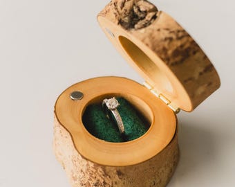 May include: A wooden ring box with a hinged lid, crafted from a natural tree branch. The interior is lined with green fabric, showcasing a diamond ring. The box is open, revealing the ring, and sits on a white surface.