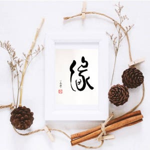May include: A white framed print with a black calligraphy character in the center. The character is surrounded by a red stamp. The print is surrounded by a white background.