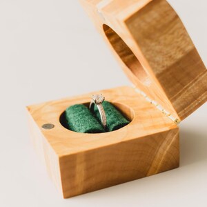 May include: A wooden ring box with a hinged lid, open to reveal a diamond engagement ring set on a green velvet cushion. The box is made of light-coloured wood and has a simple, elegant design.