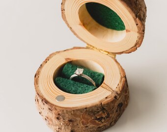 May include: A rustic wooden ring box, crafted from a tree branch, is open to reveal a diamond engagement ring. The ring rests on a bed of green fabric. The box is secured with small magnets.
