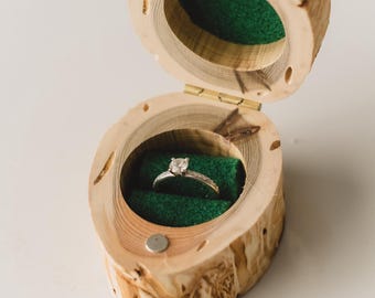 May include: A wooden ring box with a hinged lid, open to reveal a diamond ring set on a green velvet interior. The box is made from a light-colored wood with a natural, textured finish. The ring is silver with a square-cut diamond.