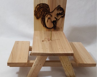 May include: A wooden squirrel feeder shaped like a picnic table with a squirrel design on the back. The feeder is made of natural wood and has a small screw in the center of the table top.