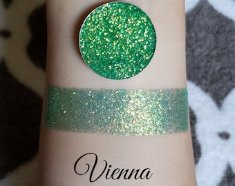 May include: A swatch of green glitter eyeshadow next to a circular eyeshadow pan. The eyeshadow is a vibrant green with gold flecks. The word "Vienna" is written in cursive below the swatch.
