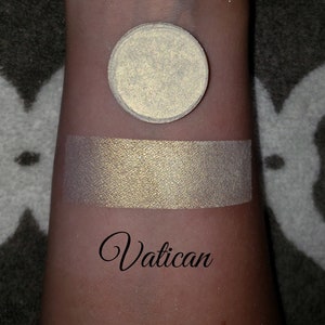 May include: A circular, pale gold eyeshadow pan and a swatch on a person's arm. The swatch and eyeshadow have a metallic finish. The word "Vatican" is written in cursive below the swatch.