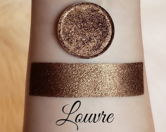 May include: A close-up of a gold eyeshadow with a shimmery finish. The eyeshadow is labeled "Louvre".