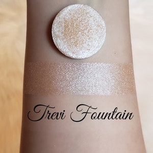 May include: A light beige eyeshadow compact and swatch on a light skin tone. The eyeshadow is round and has a textured surface. The swatch is a shimmering, light beige color. The text "Trevi Fountain" is written in cursive.