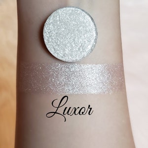 May include: A silver eyeshadow with a shimmery finish. The eyeshadow is shown in a round pan and swatched on skin. The name of the eyeshadow is "Luxor".