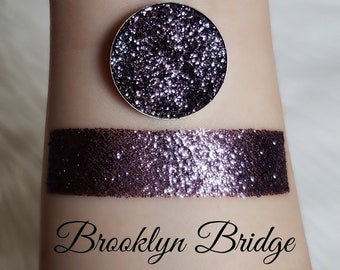 May include: A circular, silver eyeshadow pan and a swatch of glitter eyeshadow in a deep purple hue. The swatch and pan are on a light skin tone. The text "Brooklyn Bridge" is written in cursive below the swatch.