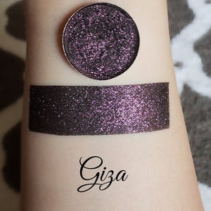 May include: A close-up of a dark purple eyeshadow with glitter, in a round metal pan, and a swatch on the arm. The swatch is rectangular. The word "Giza" is written in cursive below the swatch.