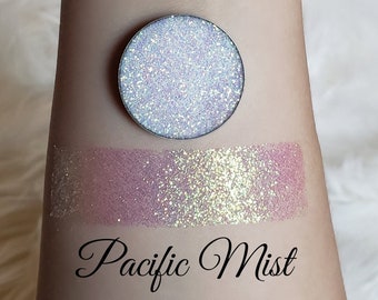 May include: A round, silver eyeshadow pan with a shimmering, light blue and silver glitter finish. Below, a swatch of the eyeshadow shows a pink and silver glitter effect. The text "Pacific Mist" is written in cursive below the swatch.