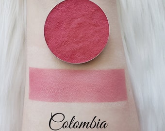 May include: A round, pressed eyeshadow in a metallic silver pan, with a swatch of the same color below. The eyeshadow and swatch are a dusty rose color. The word "Colombia" is written in cursive below the swatch.
