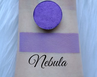 May include: A vibrant purple eyeshadow and swatch labeled "Nebula" on a light skin tone. The eyeshadow is a round compact, and the swatch is a rectangular stripe of the same color. The background is a soft, white, textured surface.