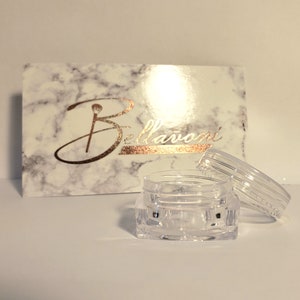 May include: A clear, small, open jar with a lid next to a marble-patterned box. The box has the word "Bellavoni" in gold script. The jar is empty and appears to be made of plastic.