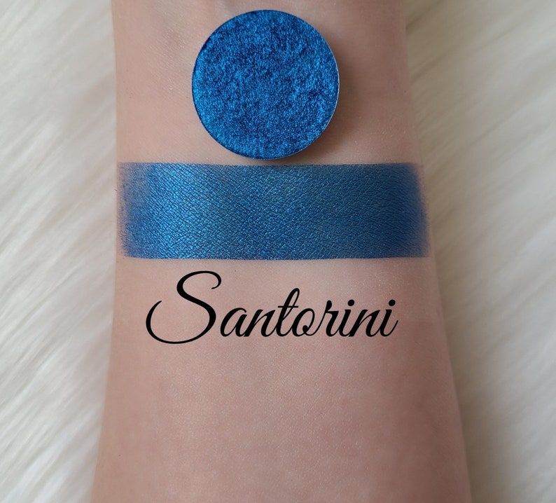 May include: A swatch of vibrant blue eyeshadow and a circular pan of the same color. The word "Santorini" is written in cursive below the swatch. The eyeshadow has a metallic finish.