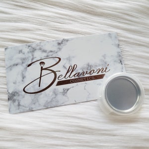 May include: A business card with a marble design and the Bellarvoni Cosmetics logo, featuring a makeup brush icon. A small, round, silver-colored makeup container with a clear plastic lid is next to the card. The background is white.