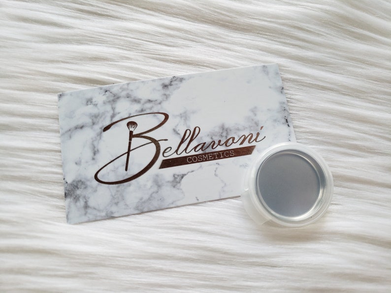 May include: A business card with a marble design and the Bellarvoni Cosmetics logo, featuring a makeup brush icon. A small, round, silver-colored makeup container with a clear plastic lid is next to the card. The background is white.