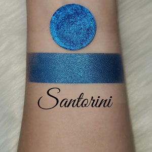 May include: A swatch of vibrant blue eyeshadow and a circular eyeshadow pan. The eyeshadow is labeled "Santorini" in a cursive font. The eyeshadow has a metallic finish and is displayed on a light skin tone.