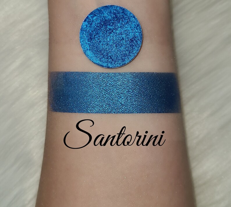 May include: A swatch of vibrant blue eyeshadow and a circular eyeshadow pan. The eyeshadow is labeled "Santorini" in a cursive font. The eyeshadow has a metallic finish and is displayed on a light skin tone.