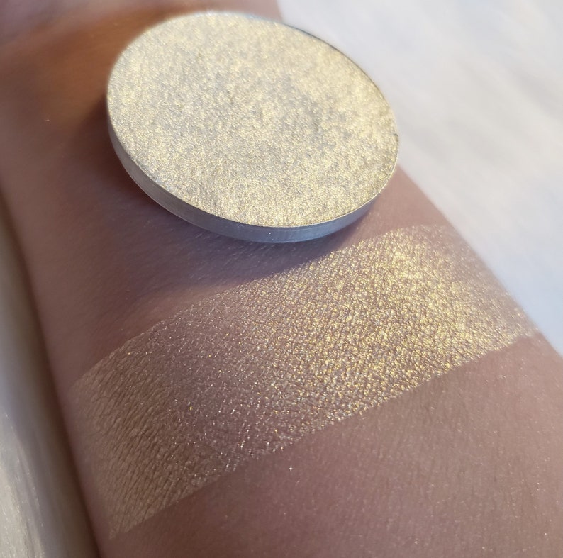 May include: A round, metallic eyeshadow pan in a light, shimmering gold color. A swatch of the same eyeshadow is applied to the arm, showing a blend of gold and silver tones. The product is designed for eye makeup.