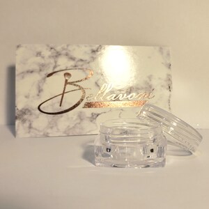 May include: A clear, small, open jar with a matching lid sits in front of a marble-patterned box. The box has the word "Bellavoni" in gold script. The jar is empty and appears to be made of plastic.