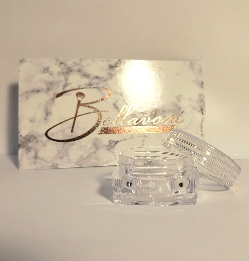 May include: A clear, small, open jar with a matching lid sits in front of a marble-patterned box. The box has the word "Bellavoni" in gold script. The jar is empty and appears to be made of plastic.