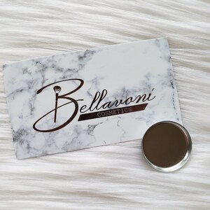 May include: A white and gray marble-patterned card with the text "Bellavoni Cosmetics" in brown script and a small, round, metallic eyeshadow pan. The card and pan are on a white, fluffy surface.