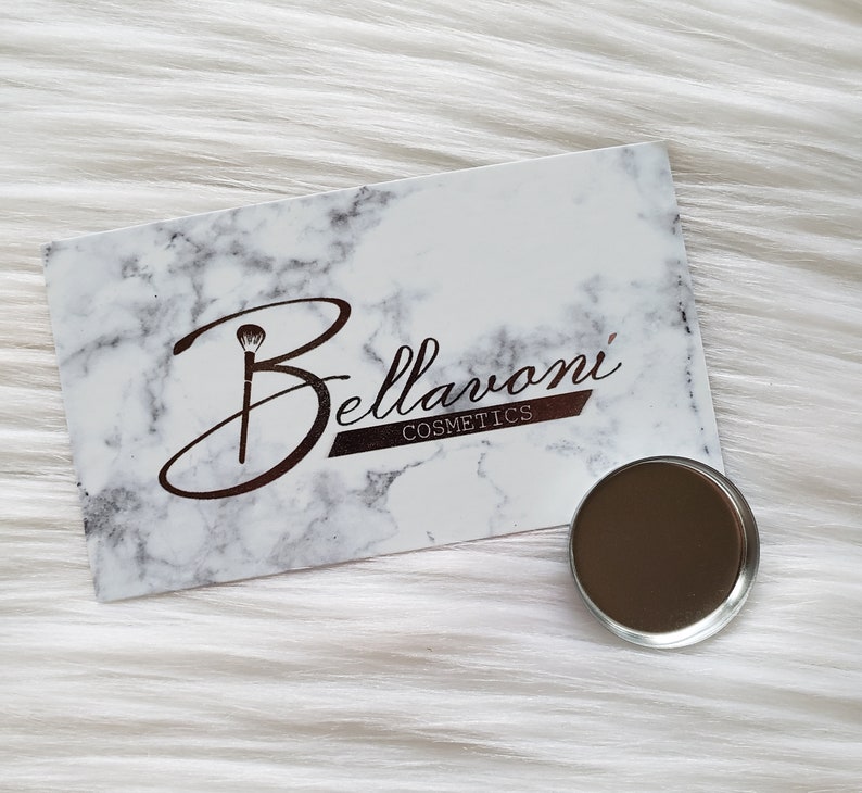 May include: A white and gray marble-patterned card with the text "Bellavoni Cosmetics" in brown script and a small, round, metallic eyeshadow pan. The card and pan are on a white, fluffy surface.