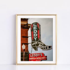 May include: Framed photograph of a vintage neon sign for Leddy's Boots & Saddlery. The sign, shaped like a cowboy boot, is black with red and green accents. The sign is mounted on a brick building.