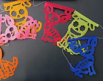 May include: A vibrant paper banner with cut-out designs. The banner showcases skull and dog skeleton shapes in orange, pink, red, green, and blue. The banner is strung together with white string, ideal for festive occasions.
