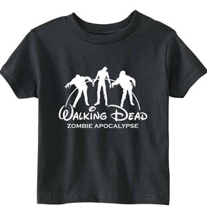 Puede incluir: Camiseta negra con un gráfico blanco de tres figuras de zombis y las palabras "Walking Dead Zombie Apocalypse" en una fuente estilizada. La camiseta es de material suave y tiene mangas cortas.
