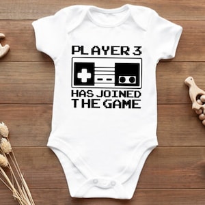 May include: White baby onesie with black text that reads "PLAYER 3 HAS JOINED THE GAME" and a black and white pixelated image of a video game controller.