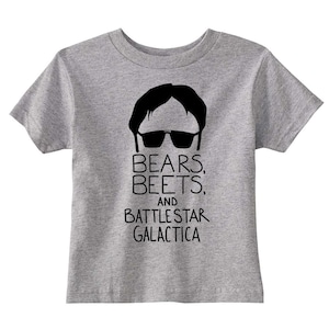 Puede incluir: Una camiseta gris para niños pequeños con una silueta negra de un hombre con gafas de sol y el texto "Bears, Beets, and Battlestar Galactica".