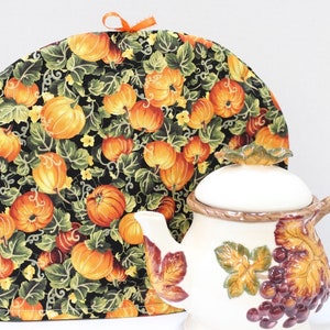 May include: A decorative tea cozy with a pumpkin and vine pattern, next to a white teapot. The cozy is black with orange pumpkins and green leaves. The teapot has a brown handle and grape design.